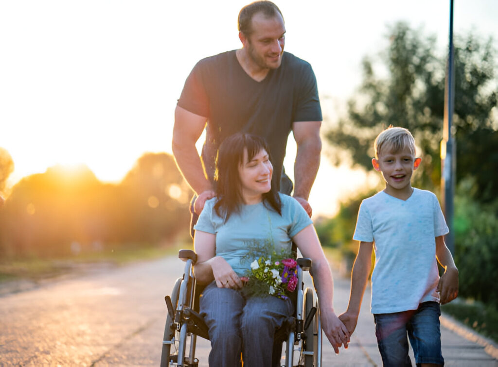 A male assists a woman in a wheelchair, working together to navigate a park pathway with a child.